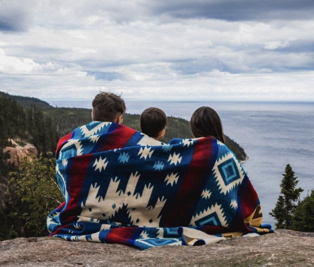 Alpakkapledd Blue Sunset 3 pers Tre personer sitter under ett pledd i naturlandskap